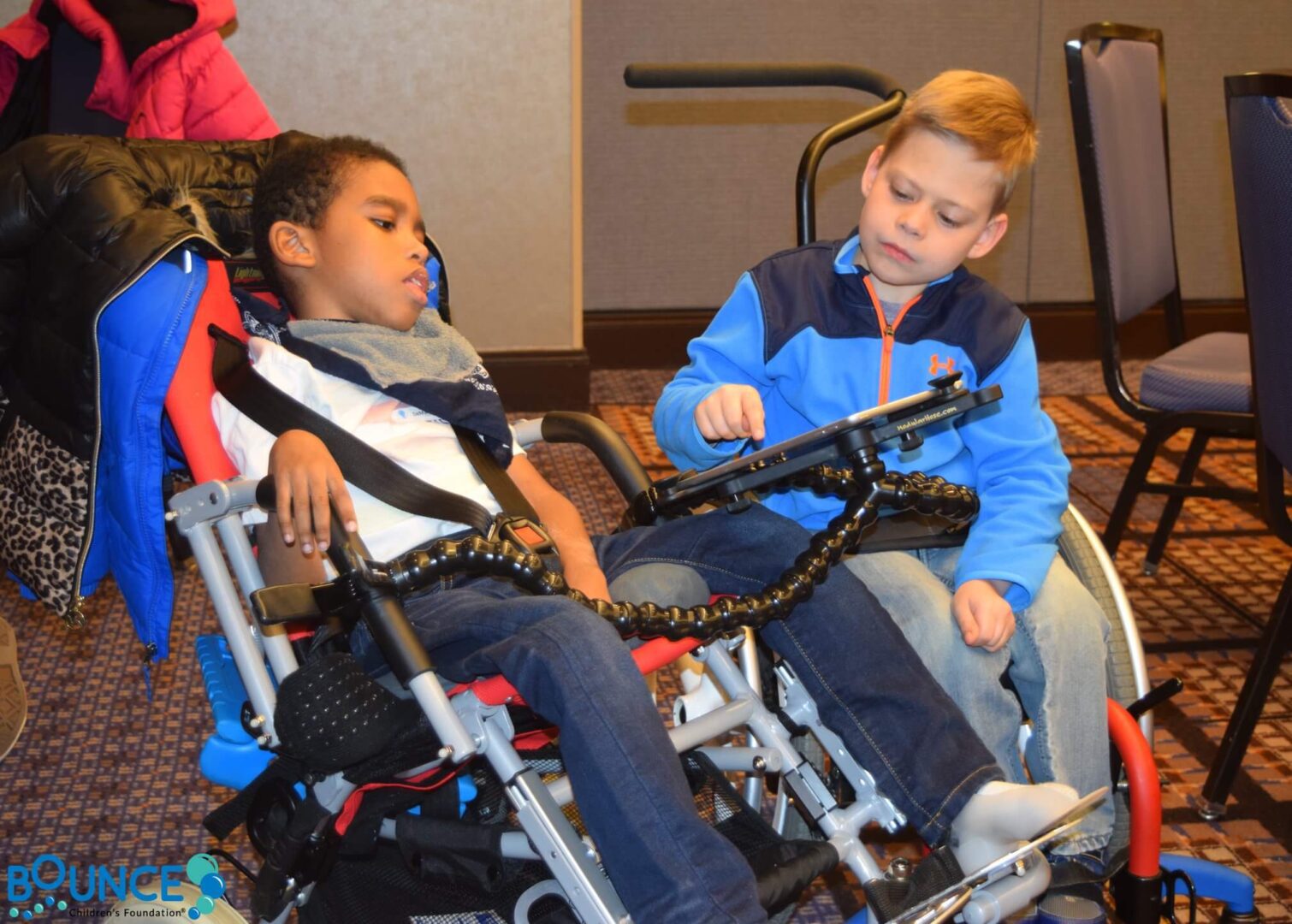 A boy helps a girl in a wheelchair with a communication device.